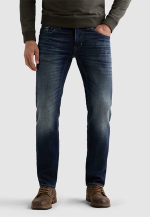 Homme portant un jean bleu foncé délavé, des bottes marron à lacets, et un sweat-shirt vert olive avec une main dans la poche.