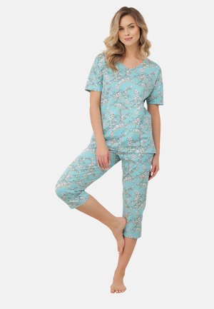 Kobieta w turkusowej bluzce w kwiaty z krótkim rękawem i spodniach do połowy łydki, stoi boso, z jedną nogą ugiętą i ręką opartą o udo.