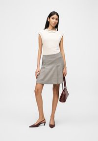 Femme portant un haut sans manches crème, une jupe à carreaux longueur genou, tenant un sac à main bordeaux et portant des talons pointus assortis, debout devant un fond blanc.