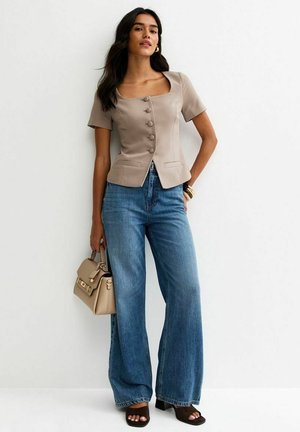 Vrouw met beige kortärmeliges, knopfverschlossenes Oberteil, weiter geschnittene blaue Jeans, schwarze offene Sandalen, die eine beige Handtasche vor weißem Hintergrund hält.