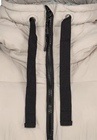 Veste matelassée beige avec une fermeture éclair noire et des cordons de serrage, présentant une texture lisse et un motif cousu. Accents métalliques remarquables inclus.