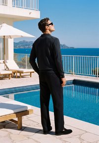 Homme portant des lunettes de soleil et des vêtements noirs se tient près d'une piscine, regardant la mer et les montagnes éloignées sous un ciel dégagé.