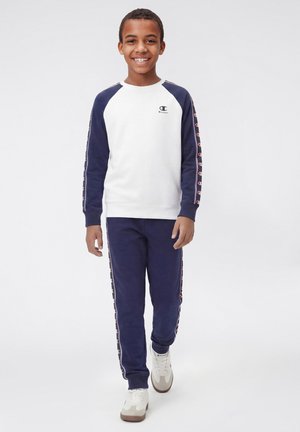 Glunderende jongen loopt naar voren en draagt een wit en marineblauw Champion sweatshirt en bijpassende marineblauwe joggingbroek met logo's op de mouwen en zijkanten van de broek.