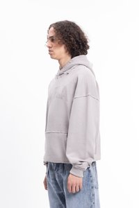 Giovane con capelli ricci che indossa un hoodie beige strappato e jeans blu larghi, in piedi di lato su uno sfondo bianco.