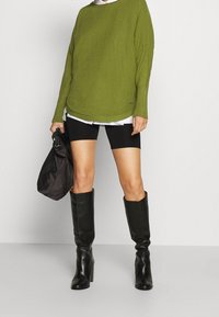Pull en maille vert olive, coupe oversize, superposé à une chemise blanche, associé à un short de vélo noir et des bottes en cuir noires jusqu'aux genoux. Sac noir.
