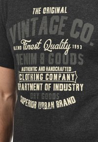 Camiseta de color gris oscuro con texto impreso en crema y gris claro, de tamaño grande y con un acabado desgastado. El diseño incluye diversas frases relacionadas con la marca y la calidad.