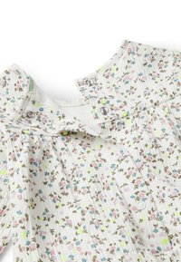 Bílý oděv s květinovým vzorem, obsahující malé zelené, růžové a modré květiny. Vyrobeno z texturované látky s patentkami u výstřihu.