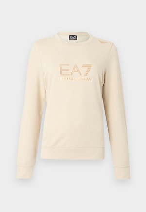 Bež majica dugih rukava od mekanog materijala, s okruglim izrezom na vratu. Na prednjem dijelu istaknuti je EA7 logo s detaljima od cirkona.