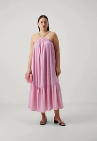 Naine, kellel on seljatagant avatud roosa halter-stiilis midi kleit, millel on volangiga ääred. Ta hoiab käes sobivat roosat klambrit ja kannab musti kõrge kontsaga sandaale.