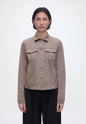 Frau trägt eine taupefarbene Jacke mit Knöpfen und Brusttaschen sowie schwarze Hosen, steht vor einem schlichten hellen Hintergrund.