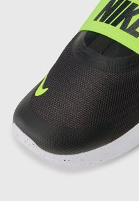 Chaussure de sport noire avec une tige en mesh et des accents verts. Présente un bout arrondi, une bande élastique et une semelle blanche mouchetée.