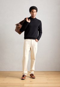 Homme debout sur un sol en bois, portant un pull noir, un pantalon beige, des chaussures marron, portant un sac en cuir marron sur l'épaule.