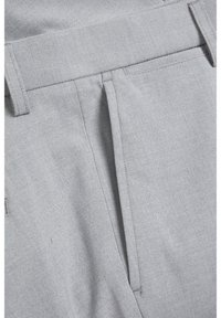 Gros plan sur un pantalon en tissu gris clair montrant la ceinture, les passants pour ceinture et un détail de poche avant plissée.