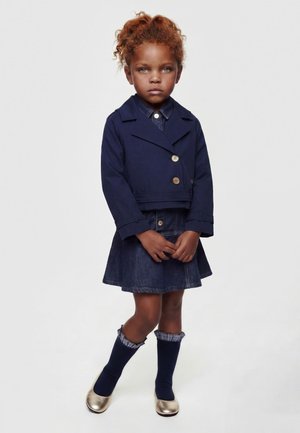 Ragazza giovane con capelli rossi ricci che indossa una giacca blu navy, una gonna di jeans, calzini blu navy con volant e scarpe dorate, in piedi davanti a uno sfondo semplice.