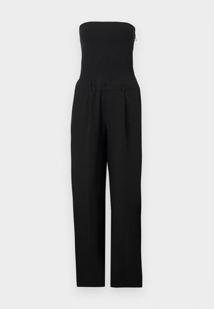 Juodas be petnešėlių kombinezonas su aptempta liemene ir platėjusiomis kelnėmis, pateiktas ant balto fono.