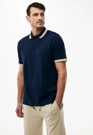 Mann trägt ein marineblaues Poloshirt mit beigem Kragen und Ärmelabschluss, beiger Hose, steht mit einer Hand in der Tasche und schaut zur Seite.