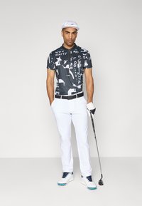 Grafisk trykt polo skjorte i svart og hvitt, kombinert med hvite bukser og golfhansker. Modellen står med en golfkølle og har på seg en hvit caps.