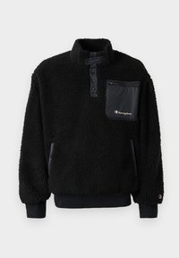 Černý fleecový pulovr s vysokým límcem, kapsou na zip na hrudi a elastickými manžetami. Obsahuje texturovanou tkaninu a hladkou část na kapsy.