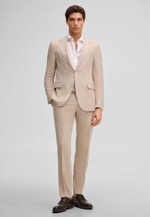 Mężczyzna ubrany w beżowy garnitur, białą koszulę i ciemnobrązowe buty wizytowe, stoi na jednolitym jasnoszarym tle.