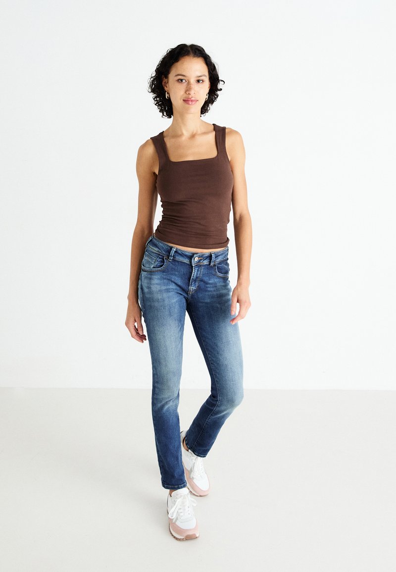 Top sin mangas marrón de escote cuadrado combinado con jeans azules de acabado desgastado y ligeras roturas, complementado con zapatillas blancas y rosas.