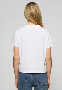 Vit kortärmad t-shirt med rund hals, gjord av mjuk bomullstyg, med en avslappnad passform och cropped fåll, sedd bakifrån.
