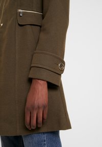 Mano de piel oscura descansando al lado de un abrigo verde oliva con puño abotonado y bolsillo con cremallera, combinado con unos pantalones vaqueros azules.