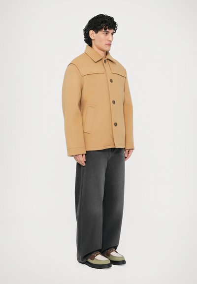 Ταμπά μάλλινο σακάκι με γιακά, κουμπιά μπροστά, δύο πλαϊνές τσέπες και καφέ κουμπιά· συνδυασμένο με φαρδιά σκούρα τζιν και πολύχρωμα παπούτσια.