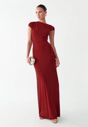 Vrouw draagt een vloerlange aansluitende rode jurk met korte mouwen en een zijkant ruches, houdt een kleine gouden clutch vast en draagt gouden sandalen.