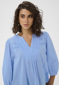 Blouse bleu clair avec un col en V, des détails plissés et des accents texturés. Comprend des manches ballon et une coupe décontractée.