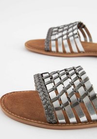 Sandales métalliques en argent tressé avec une sangle grise texturée, semelle en daim marron et semelle plate. Détails de couture subtils autour des bords.