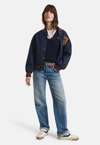 Marineblau gestreifte Bomberjacke mit orangefarbenen Akzenten, getragen über einem marineblauen Kabelstrickpullover. Hellblaue Jeans und weiße Sneaker runden das Outfit ab.