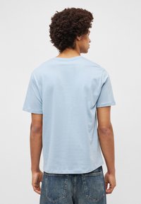 Ανοιχτό μπλε βαμβακερό μπλουζάκι με χαλαρή εφαρμογή, στρογγυλή λαιμόκοψη και κοντά μανίκια. Από την πίσω πλευρά φαίνεται υφή λεία και μινιμαλιστική σχεδίαση.