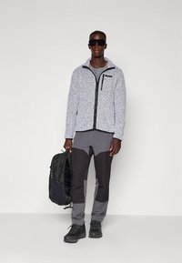 Graue Fleecejacke mit schwarzem Reißverschluss und Akzenten, kombiniert mit grauer und schwarzer Hose. Das Model hält einen schwarzen Rucksack und trägt dunkle Sonnenbrillen.