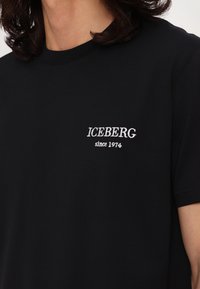 T-shirt noir avec texte blanc "ICEBERG since 1974" sur le côté gauche de la poitrine, porté par une personne aux cheveux ondulés foncés partiellement visibles.