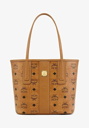 Tan leather tote bag with dual handles, featuring black MCM logo patterns and diamond accents. Gold hardware detail in the center.