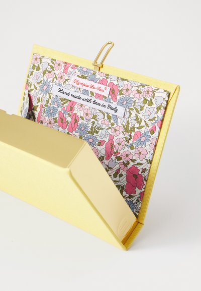 Clutch în formă de carte aurie, căptușit cu țesătură florală. Dispune de un închizător aurit. Textul spune "Fabricat manual cu drag în Italia" și eticheta de brand.