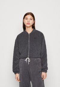 Jonge vrouw met steil bruin haar, gekleed in een donkergijze gewatteerde zip-up jack en bijpassende sweats met een witte trekkoord tegen een effen achtergrond.