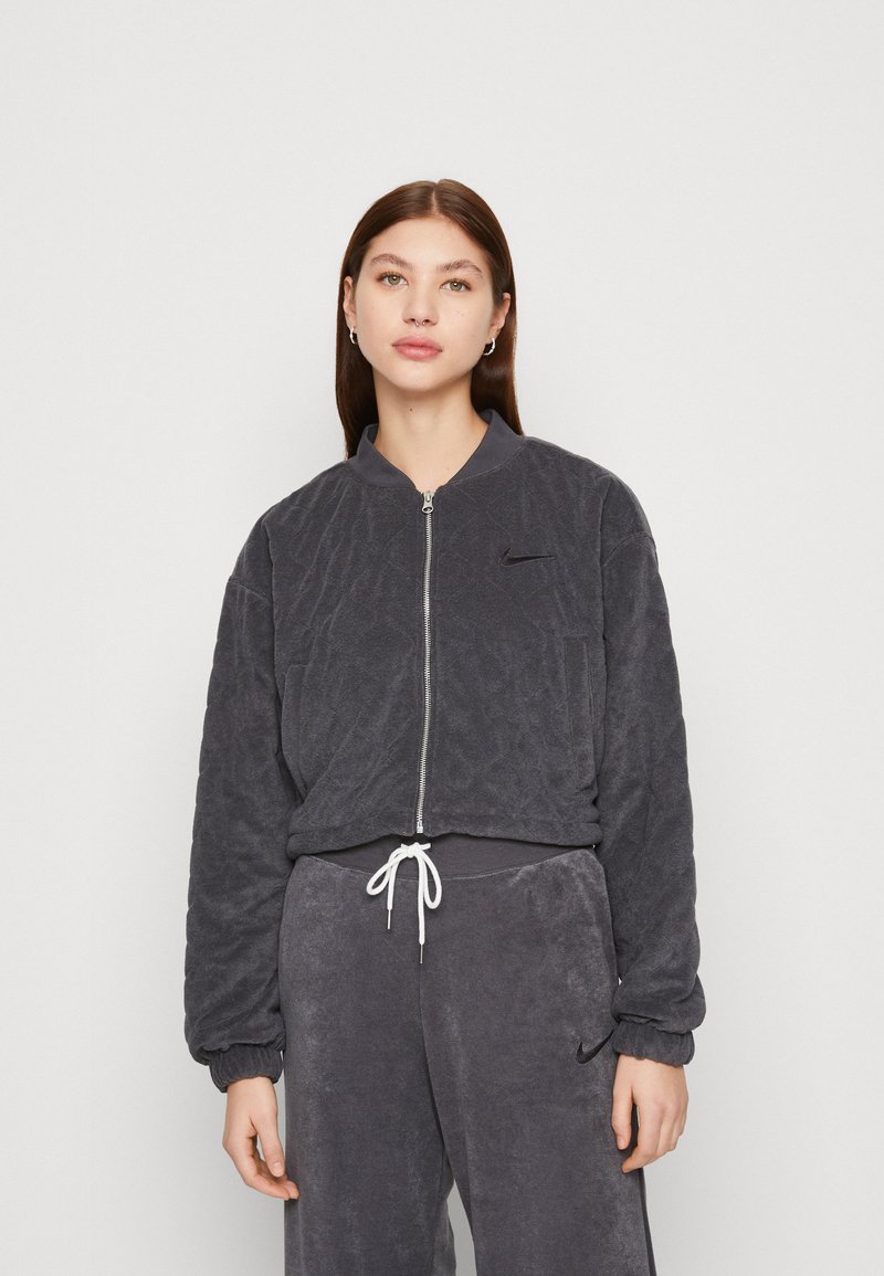 Jonge vrouw met steil bruin haar, gekleed in een donkergijze gewatteerde zip-up jack en bijpassende sweats met een witte trekkoord tegen een effen achtergrond.