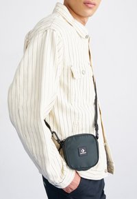 Kwadratowa zielona torba na ramię z materiału, z czarną naszywką z logo. Noszona z kremową marynarką w paski, zabezpieczona czarnym paskiem.