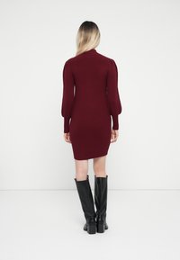 Burgundy knit dress with long puff sleeves and a high neck. Worn with knee-high black leather boots, featuring a block heel.