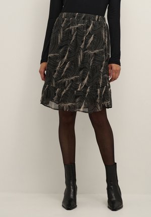 Femme portant une jupe noire à motifs longueur genou avec des collants noirs et des bottines noires, debout devant un fond uni.
