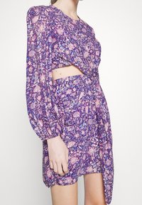 Květinové krátké šaty v purpurové barvě s růžovými a modrými akcenty. Mají výřez, dlouhé balónové rukávy a řasený pas.