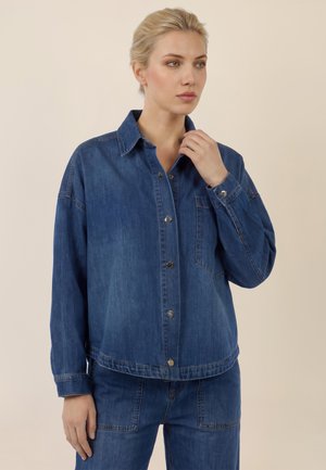 Blonde woman wearing a blue denim jacket and matching pants, standing against a beige background, looking slightly to the side.
