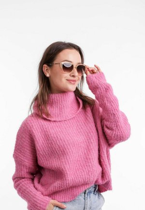 Růžový pletený svetr s vysokým límcem a žebrovanou texturou, doplněný slunečními brýlemi se zlatými akcenty. Modelka nosí džíny šortky.