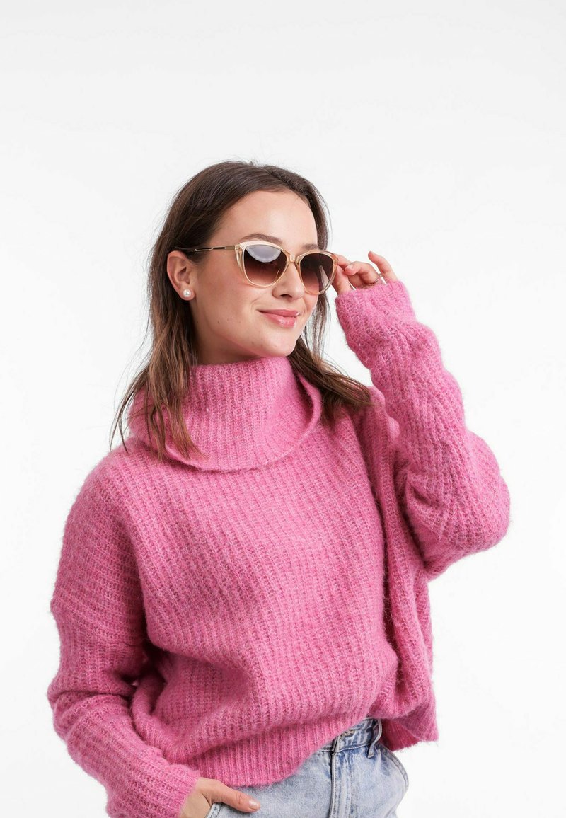 Růžový pletený svetr s vysokým límcem a žebrovanou texturou, doplněný slunečními brýlemi se zlatými akcenty. Modelka nosí džíny šortky.