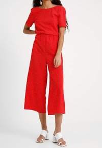 Röd jumpsuit med korta ärmar, draperad midja, breda cropped ben och vita prickar. Tillsammans med vita blockklack sandaler.