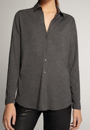 Femme portant une chemise à manches longues gris foncé avec boutons, col et poche poitrine, associée à un pantalon noir.