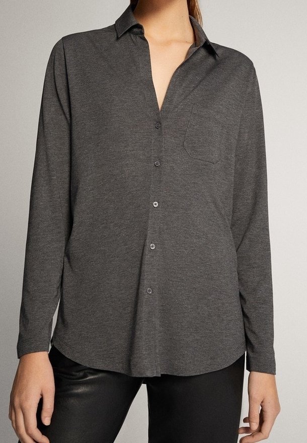 Femme portant une chemise à manches longues gris foncé avec boutons, col et poche poitrine, associée à un pantalon noir.