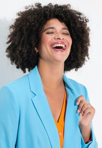 Blazer azzurro chiaro con rever a punta sopra una maglia arancione. Capelli ricci scuri. Sfondo luminoso, espressione sorridente e mano appoggiata sul colletto.