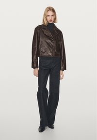Eine Frau in einer dunkelbraunen Lederjacke, einem schwarzen Rollkragenpullover, dunklen weiten Jeans und schwarzen spitzen Schuhen steht vor einem schlichten Hintergrund.